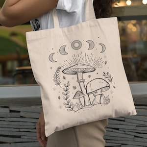 K&ouml;nnte beinhalten: Eine beige Canvas-Tragetasche mit einer schwarz-wei&szlig;en Illustration von Pilzen, Kristallen und Monden. Die Illustration ist von kleinen Sternen und Bl&auml;ttern umgeben.