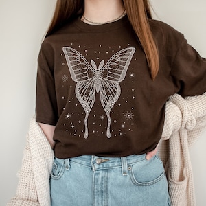 May include: A brown t-shirt with a white outline of a moth with stars and a celestial theme. The shirt is being worn by a person with a brown cardigan.