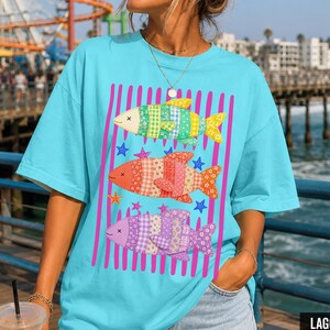 May include: A turquoise Comfort Colors t-shirt featuring a fish design. The design shows three fish with patchwork patterns in green, orange, and purple, set against pink vertical stripes. The shirt is labelled "Lagoon Blue".