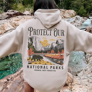 Può includere: Una felpa con cappuccio beige con una grafica sul retro che recita "PROTECT OUR NATIONAL PARKS" con un'illustrazione di un orso, montagne e alberi. Il testo "PARKS, NOT PROFITS" è sotto l'illustrazione. La felpa con cappuccio viene indossata all'aperto.