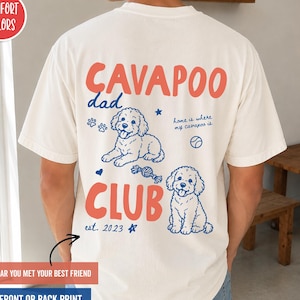 May include: An ivory short-sleeve t-shirt featuring a back graphic with the red text CAVAPOO CLUB and blue line art of two dogs, paw prints, a ball, and the text dad, home is where my cavapoo is, and est. 2023. A red circle label reads COMFORT COLORS.
