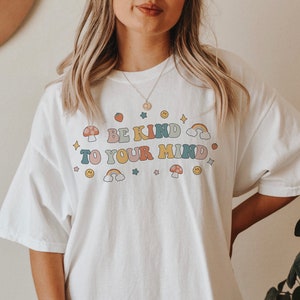 Puede incluir: Camiseta blanca de gran tama&ntilde;o con un estampado gr&aacute;fico del texto "Be Kind To Your Mind" en letras de colores con detalles de arco&iacute;ris, setas y estrellas.