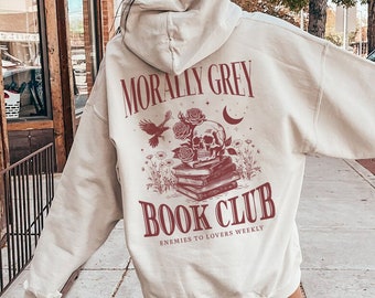 May include: A cream-colored hoodie with a graphic print of a skull, books, and flowers. The text "Morally Grey Book Club" is printed in red on the hoodie. The text "Enemies to Lovers Weekly" is printed in smaller red letters below the main text.