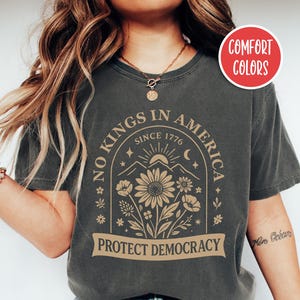 May include: Dark grey t-shirt with the text "NO KINGS IN AMERICA SINCE 1776 PROTECT DEMOCRACY". The shirt features a floral design with a mountain and sun graphic. The shirt is a comfort color.