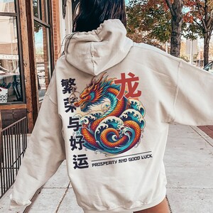 May include: A cream-colored hoodie with a large graphic print of a red dragon with blue and orange waves. The text "Prosperity and Good Luck" is printed below the dragon. The Chinese characters "繁荣与好运" are printed on the left side of the dragon.