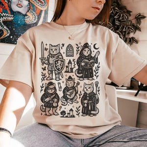 May include: A beige t-shirt with a black and white illustration of five cats dressed as fantasy characters. The cats are a knight, a wizard, a bard, a rogue, and a cleric. The illustration is surrounded by stars, leaves, and a castle.