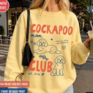 Könnte beinhalten: Ein blassgelbes Sweatshirt mit Rundhalsausschnitt, rotem Schriftzug COCKAPOO MOM CLUB und blauem Text est. 2026. Das Design zeigt Illustrationen von zwei Cockapoo-Hunden, einem Ball, einem Spielzeug und dem Satz home is where my cockapoo is.
