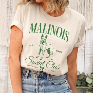 May include: Cream-colored t-shirt featuring the text "Malinois Social Club" in green, with a graphic of a dog. The text includes "Est. 1991" and "Santa Rosa, California". The shirt is paired with blue jeans.