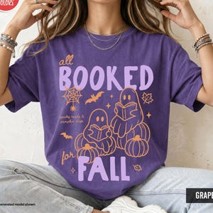 以下が含まれることがあります： 「all BOOKED for FALL」および「spooky reads & pumpkin days」という文字がプリントされたグレープパープルのTシャツ。デザインには、本を読む2体のゴースト、カボチャ、クモの巣、コウモリ、オレンジとライトパープルの葉が描かれています。