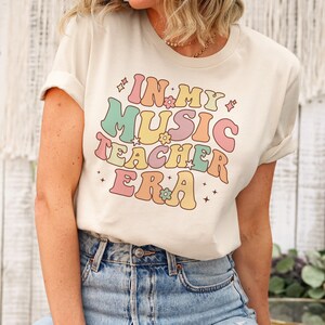 以下が含まれることがあります： レトロなフォントでカラフルな文字と花で「In My Music Teacher Era」と書かれた薄いベージュのTシャツを着た女性。彼女は、ダメージ加工が施されたライトブルーのジーンズを履いています。
