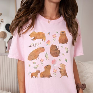 May include: A pink oversized t-shirt with a graphic print of multiple capybaras in a forest setting. The capybaras are brown and are depicted in various poses, including sitting, standing, and walking. The forest setting includes green leaves, pink flowers, and a mushroom.