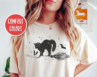 May include: A cream coloured t-shirt featuring a black cat riding a broomstick with bats flying around it. The shirt has the words "Comfort Colors" on a red circle.