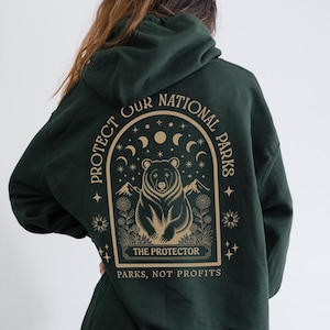 Può includere: Felpa con cappuccio verde scuro con una grafica sul retro. La grafica presenta un orso stilizzato circondato dalla scritta "PROTECT OUR NATIONAL PARKS", con le parole "THE PROTECTOR" e "PARKS, NOT PROFITS" sottostanti.