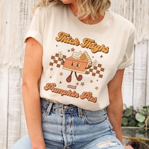 May include: A white t-shirt with a retro-style graphic of a smiling pumpkin pie slice with the text "Thick Thighs and Pumpkin Pies".