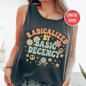 May include: A black tank top with the text "RADICALIZED BY BASIC DECENCY" in a retro font with flower and peace sign graphics. The shirt is a comfort color and is pepper colored.