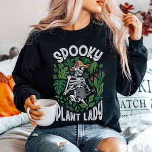 May include: Black sweatshirt with a spooky plant lady graphic. The graphic features a skeleton holding a watering can in a garden with green leaves and flowers. The text "Spooky Plant Lady" is printed below the graphic.