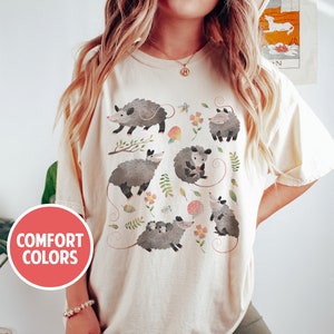 May include: A beige t-shirt with a graphic print of multiple opossums in a woodland setting. The opossums are depicted in a cartoon style with floral accents. The text "COMFORT COLORS" is visible in a red circle on the left side of the image.
