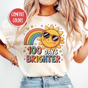 May include: A white t-shirt featuring a rainbow and a smiling sun wearing sunglasses. The text "100 Days Brighter" is printed on the shirt.