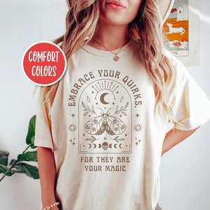 May include: A beige t-shirt with a brown graphic design featuring a moth, crescent moon, stars, and the text "Embrace Your Quirks. For They Are Your Magic."