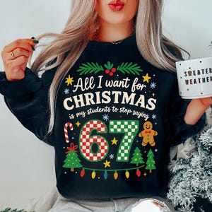 May include: Black sweatshirt with the text "All I want for CHRISTMAS is my students to stop saying" and the number "67". The design includes Christmas trees, a gingerbread man, and a candy cane. A white mug with the text "SWEATER WEATHER" is held in the hand.