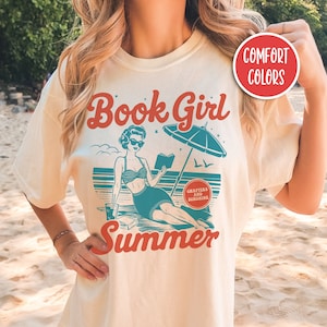 K&ouml;nnte beinhalten: Ein wei&szlig;es T-Shirt mit einer Vintage-Illustration einer Frau, die am Strand ein Buch liest. Der Text "Book Girl Summer" ist auf dem Shirt gedruckt, zusammen mit dem Satz "Chapters and Sunshine".