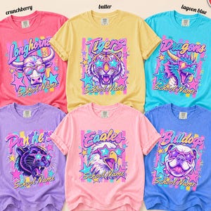 May include: Six short-sleeve t-shirts in crunchberry, butter, lagoon blue, violet, blossom, and flo blue. Each shirt features a graphic of an animal wearing star-shaped glasses, colorful stripes, stars, and the text School Name and team names.