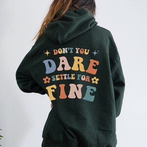 Puede incluir: Una sudadera con capucha verde oscuro con el texto "Don't you dare settle for fine" en letras de colores en la espalda.