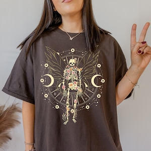 May include: A dark brown t-shirt with a floral skeleton design. The skeleton is surrounded by a circle of stars and moons. The design is in shades of gold, white, and pink.