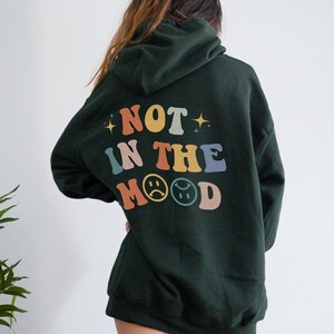 Pu&ograve; includere: Una felpa con cappuccio verde scuro con il testo "NOT IN THE MOOD" in colori arcobaleno con emoji di facce tristi.