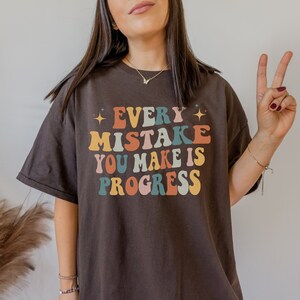 May include: A dark brown t-shirt with the text "EVERY MISTAKE YOU MAKE IS PROGRESS" in colorful, retro-style lettering. The design includes stars and a vintage font, creating a motivational and positive message.