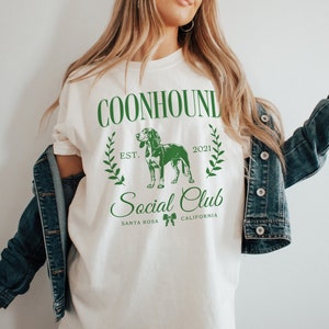 May include: Cream-colored t-shirt with a green graphic. The graphic includes the text "COONHOUND Social Club" with a dog illustration and the words "SANTA ROSA CALIFORNIA". The shirt has a relaxed fit and short sleeves.