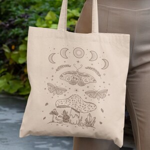 May include: A white tote bag with a brown design of a mushroom, moths, and crescent moons. The bag is being held by a person wearing a white t-shirt with the text "You are made of magic" and a pink and purple tiger design.