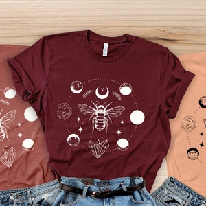 May include: Three t-shirts with a white bee and moon design. The shirts are in maroon, peach, and light brown.