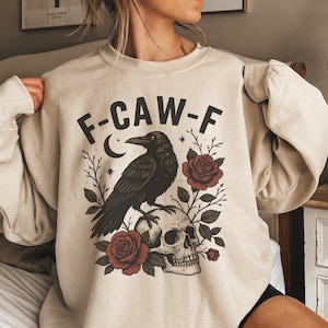Puede incluir: Sudadera beige con un diseño de un cuervo negro posado sobre una calavera, rodeado de rosas y follaje. El texto "F-CAW-F" está encima del cuervo. El diseño tiene una estética vintage y gótica.