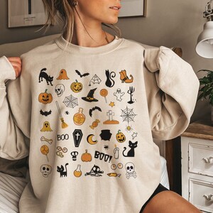 May include: A beige sweatshirt with a Halloween-themed design featuring various icons such as pumpkins, ghosts, bats, skulls, and the words "BOO" and "BOO!"