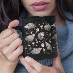 May include: A black ceramic mug with a white handle features a repeating pattern of opossums, mushrooms, and flowers.