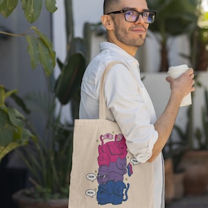 K&ouml;nnte beinhalten: Eine beige Canvas-Tragetasche mit einer grafischen Illustration eines vertikalen Stapels aus vier Katzen in Rosa-, Lila- und Blaut&ouml;nen. Die Katzen haben Sprechblasen mit dem Text "Them" und "Meow", und eine Katze h&auml;lt eine kleine Pride-Flagge.