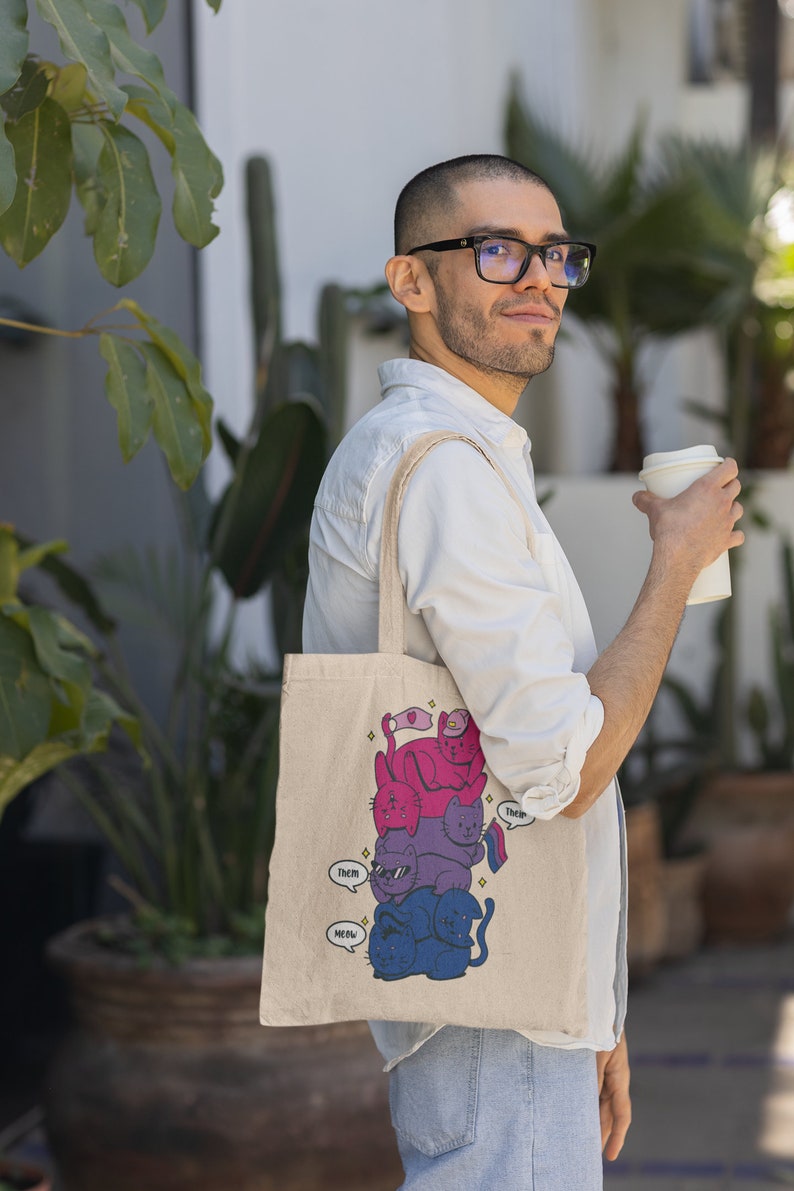 K&ouml;nnte beinhalten: Eine beige Canvas-Tragetasche mit einer grafischen Illustration eines vertikalen Stapels aus vier Katzen in Rosa-, Lila- und Blaut&ouml;nen. Die Katzen haben Sprechblasen mit dem Text "Them" und "Meow", und eine Katze h&auml;lt eine kleine Pride-Flagge.