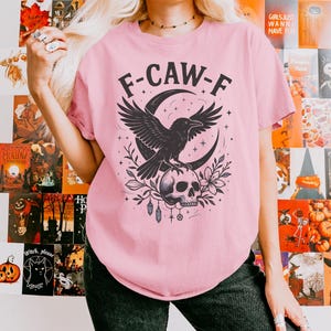 May include: A light pink t-shirt featuring a black graphic of a raven perched on a skull, with a crescent moon and floral accents. The text "F-CAW-F" is above the raven. The shirt is part of a Halloween or gothic-themed collection.