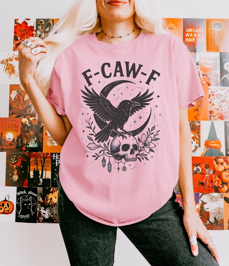May include: A light pink t-shirt featuring a black graphic of a raven perched on a skull, with a crescent moon and floral accents. The text "F-CAW-F" is above the raven. The shirt is part of a Halloween or gothic-themed collection.