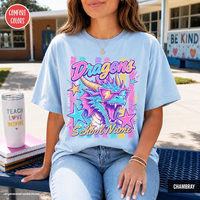 May include: A light blue t-shirt featuring a graphic of a blue dragon wearing pink sunglasses, surrounded by colorful stars and vertical stripes. The text reads Dragons and School Name in a stylized font. The shirt has a crew neck and short sleeves.