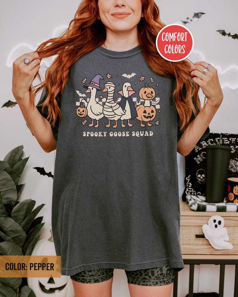 May include: Dark grey t-shirt with a Halloween graphic. The design shows cartoon geese dressed as a witch, mummy, vampire, and pumpkin. The text "Spooky Goose Squad" is below the graphic. The shirt is labelled "Comfort Colors" and the colour is "Pepper".