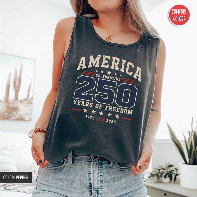 以下が含まれることがあります： 「AMERICA CELEBRATING 250 YEARS OF FREEDOM 1776-2026」の文字が白とネイビーでプリントされた、濃いグレーのタンクトップ。 タンクトップはノースリーブで、柔らかい素材で作られています。色はペッパーです。