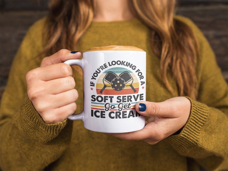 May include: White ceramic mug with a retro-style design featuring a pickleball paddle and ball. The text on the mug reads "If you're looking for a soft serve go get ice cream."