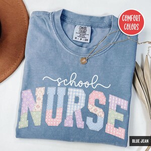 Könnte beinhalten: Ein blaues Comfort Colors T-Shirt mit den Worten "school NURSE" in einem Patchwork-Design. Das Shirt hat einen Rundhalsausschnitt und kurze Ärmel. Eine goldene Halskette mit Anhänger ist über dem Shirt drapiert.