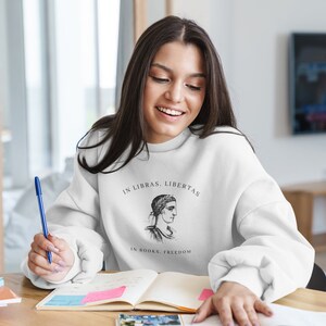 May include: A white sweatshirt with a black and white illustration of a woman's head with a laurel wreath. The text "In Libras, Libertas" and "In Books, Freedom" is printed on the sweatshirt.