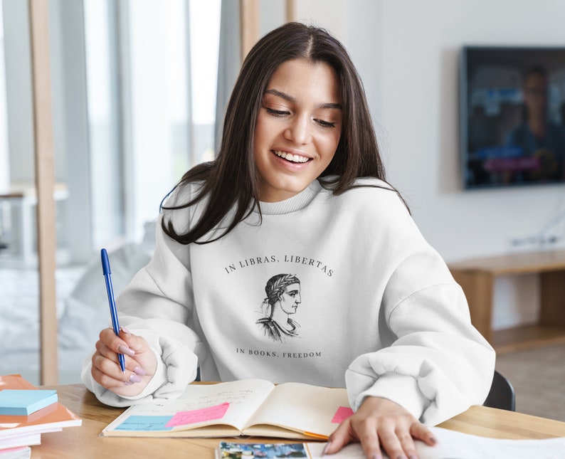May include: A white sweatshirt with a black and white illustration of a woman's head with a laurel wreath. The text "In Libras, Libertas" and "In Books, Freedom" is printed on the sweatshirt.