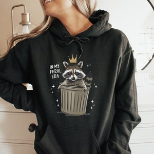 May include: Black hoodie featuring a graphic of a raccoon wearing a crown and sitting in a bin. The text "In My Feral Era" is printed on the hoodie.