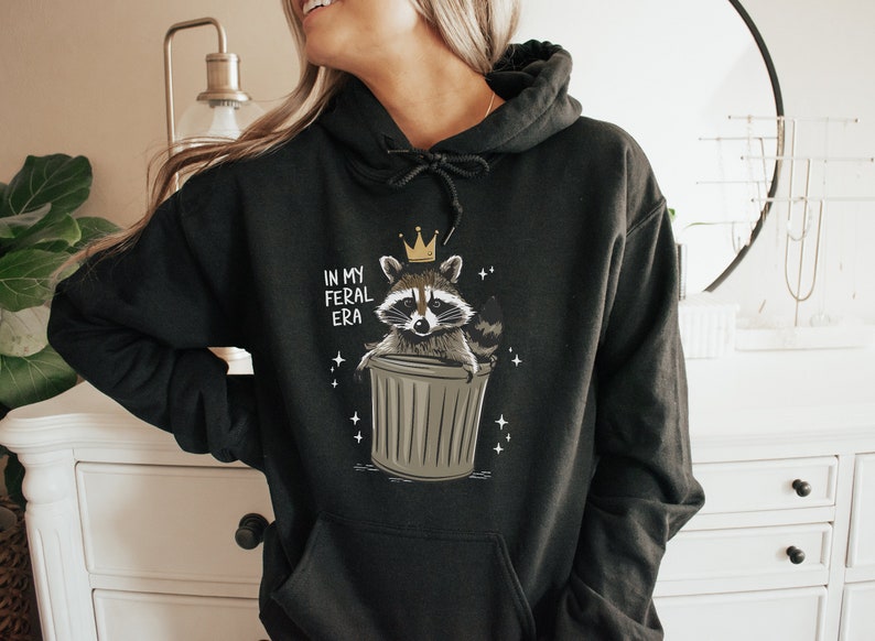 May include: Black hoodie featuring a graphic of a raccoon wearing a crown and sitting in a bin. The text "In My Feral Era" is printed on the hoodie.