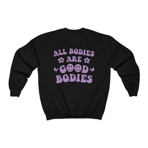 May include: Black sweatshirt featuring the lavender text "ALL BODIES ARE GOOD BODIES". The design incorporates flowers, smiley faces, and butterflies. A relaxed, casual top for everyday wear.